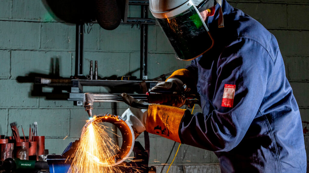 Worker in full protective gear using a cutting torch on a metal pipe, with bright sparks flying in a workshop environment.
