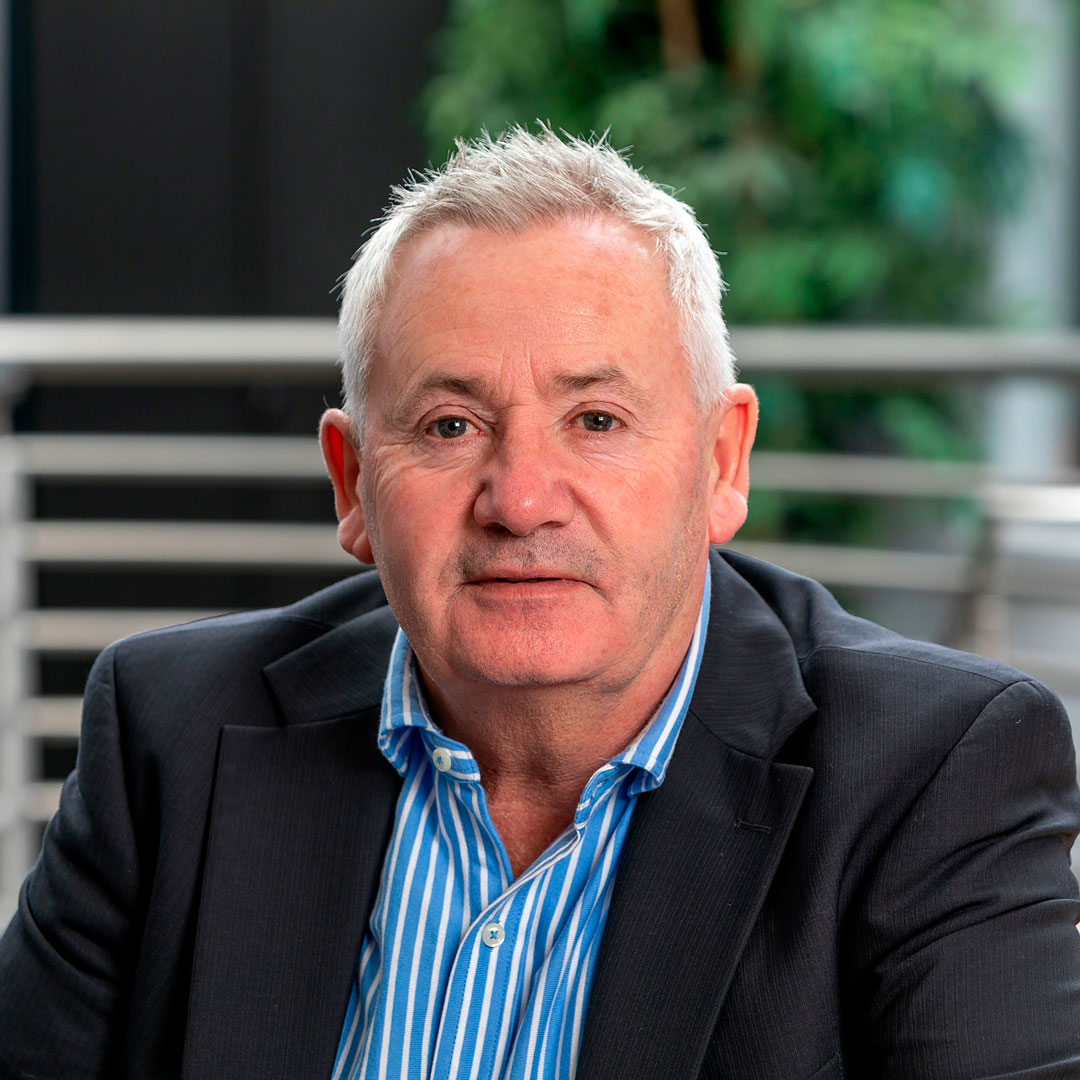 Man with grey hair wearing a striped blue and white shirt and a black blazer.